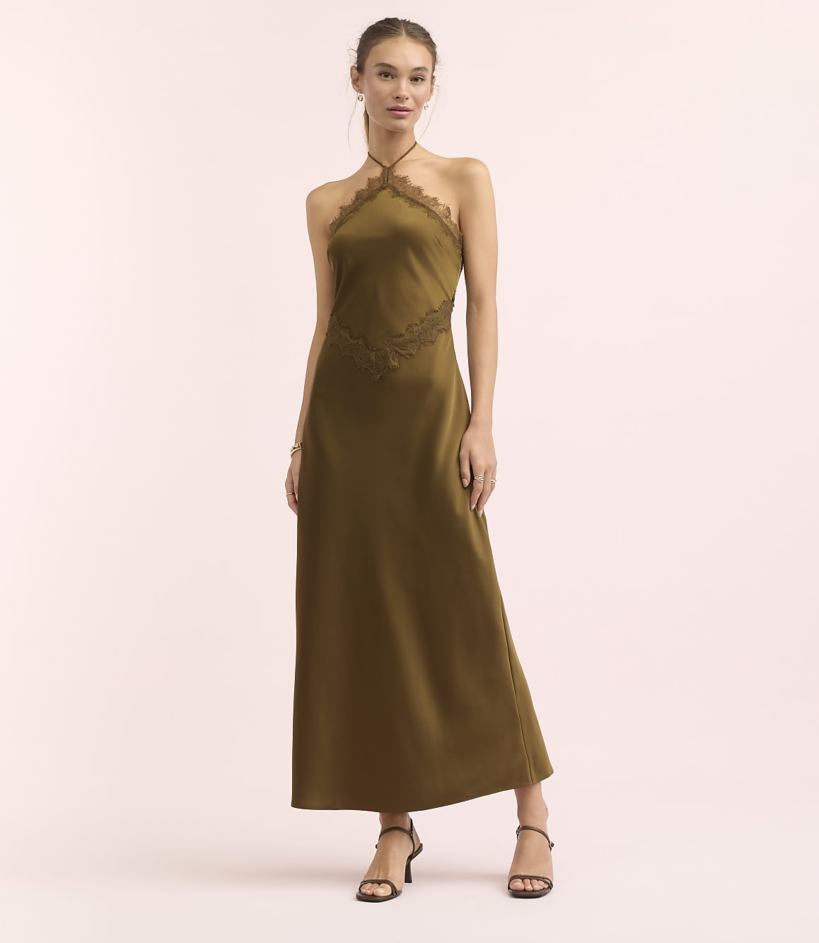 A woman in a long, olive-green halter dress with lace detailing stands against a soft pink background.