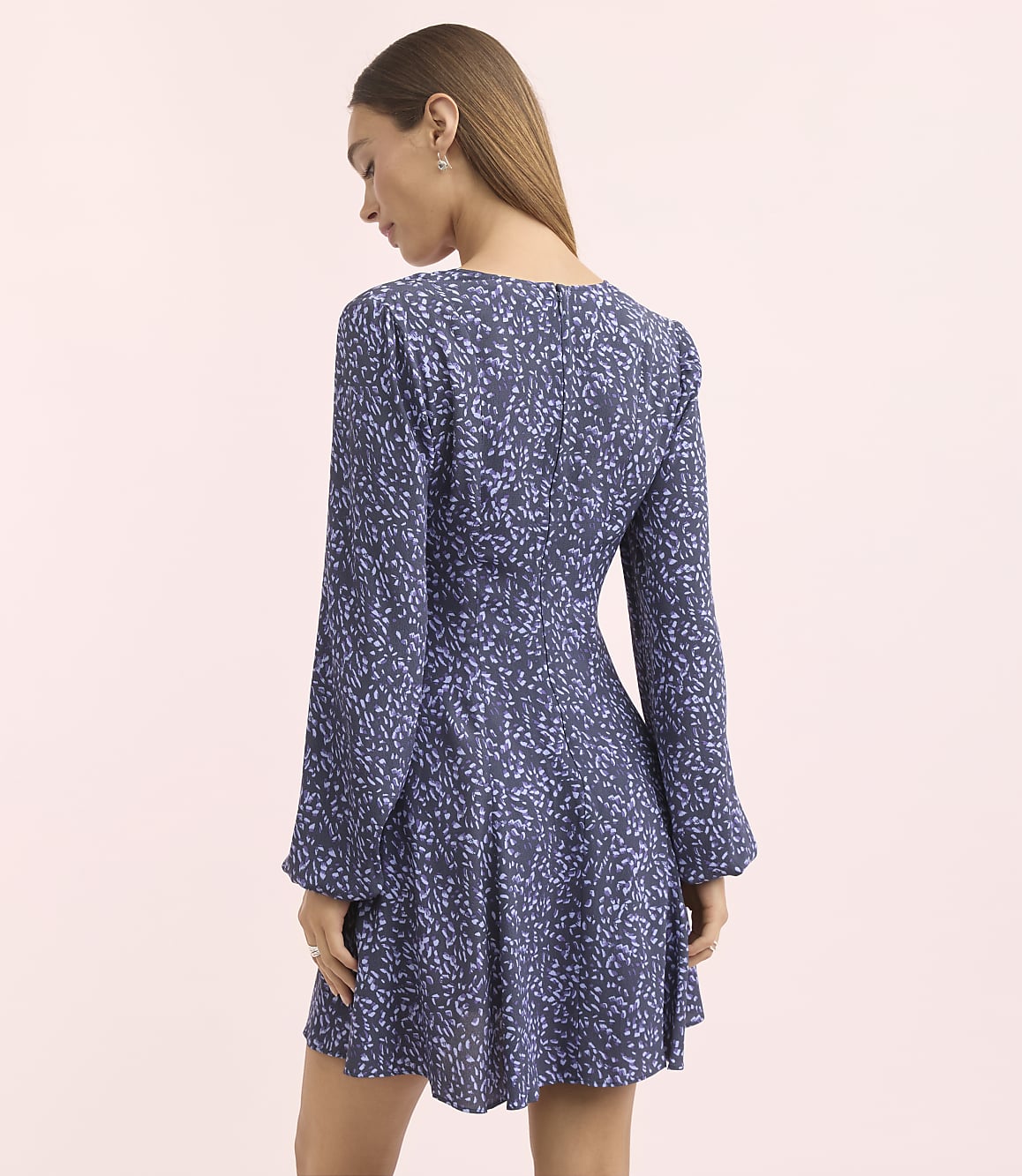 A woman in a blue floral dress stands against a plain pink background.