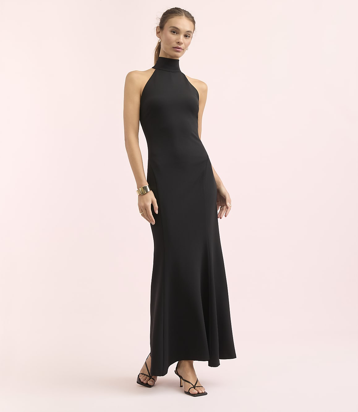 A woman in a black halter neck gown stands against a soft pink background.