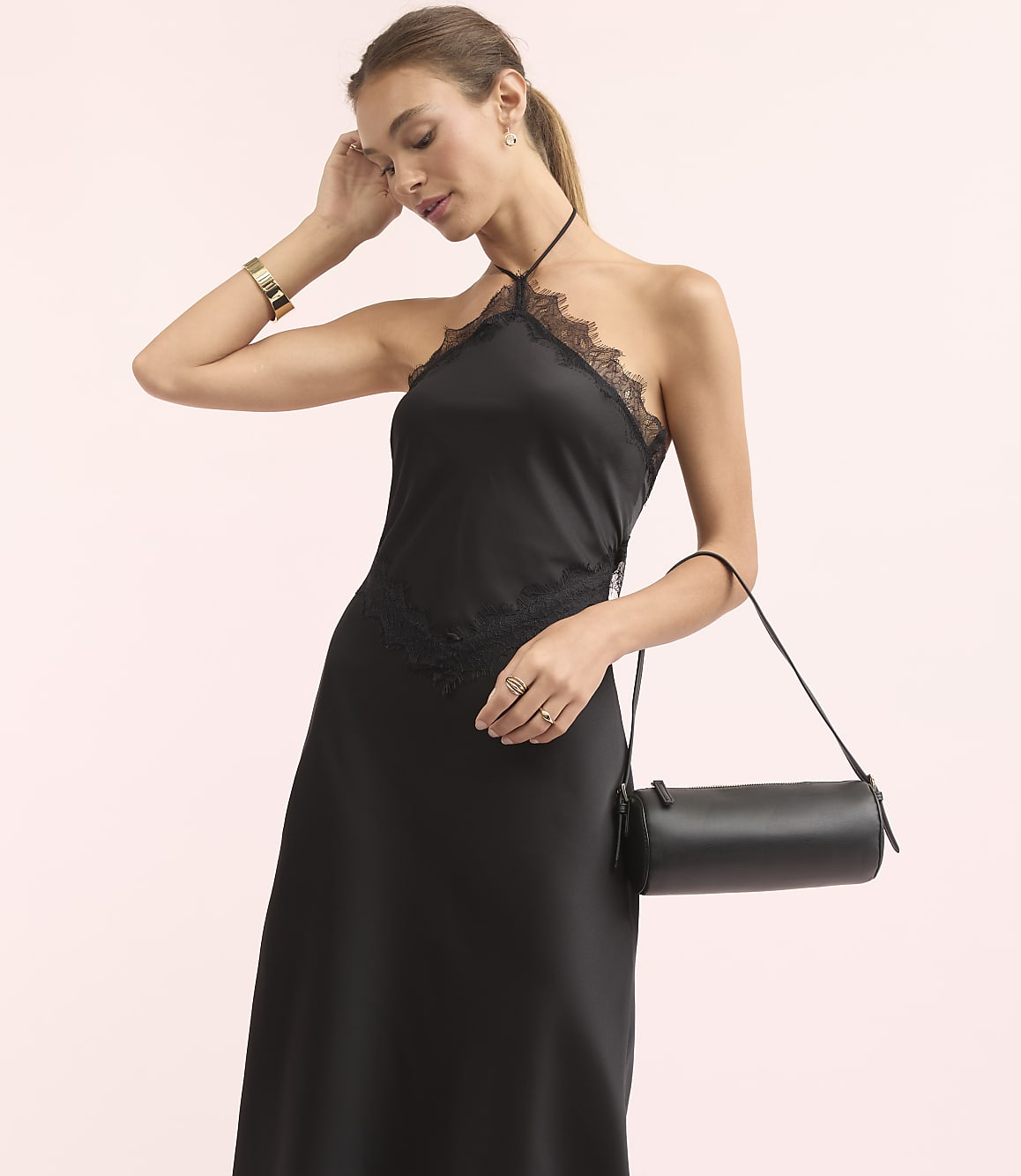 A woman in a black lace-trimmed halter dress and gold bracelet holds a cylindrical black purse against a soft pink background.