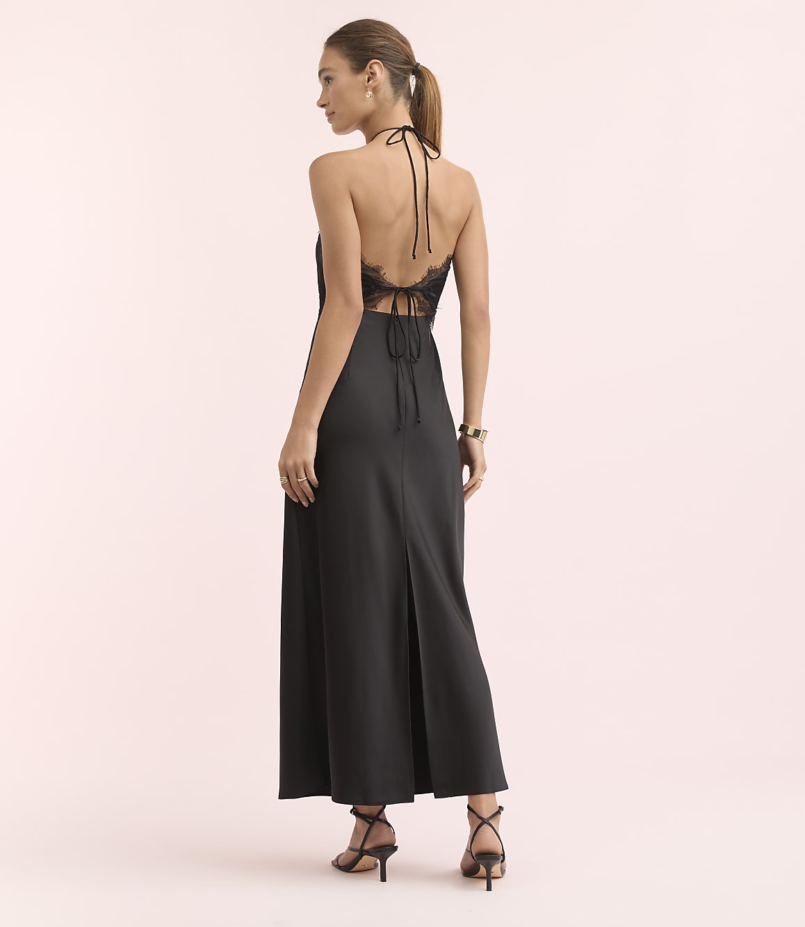 A woman in a black halter dress with lace detailing stands against a soft pink background.