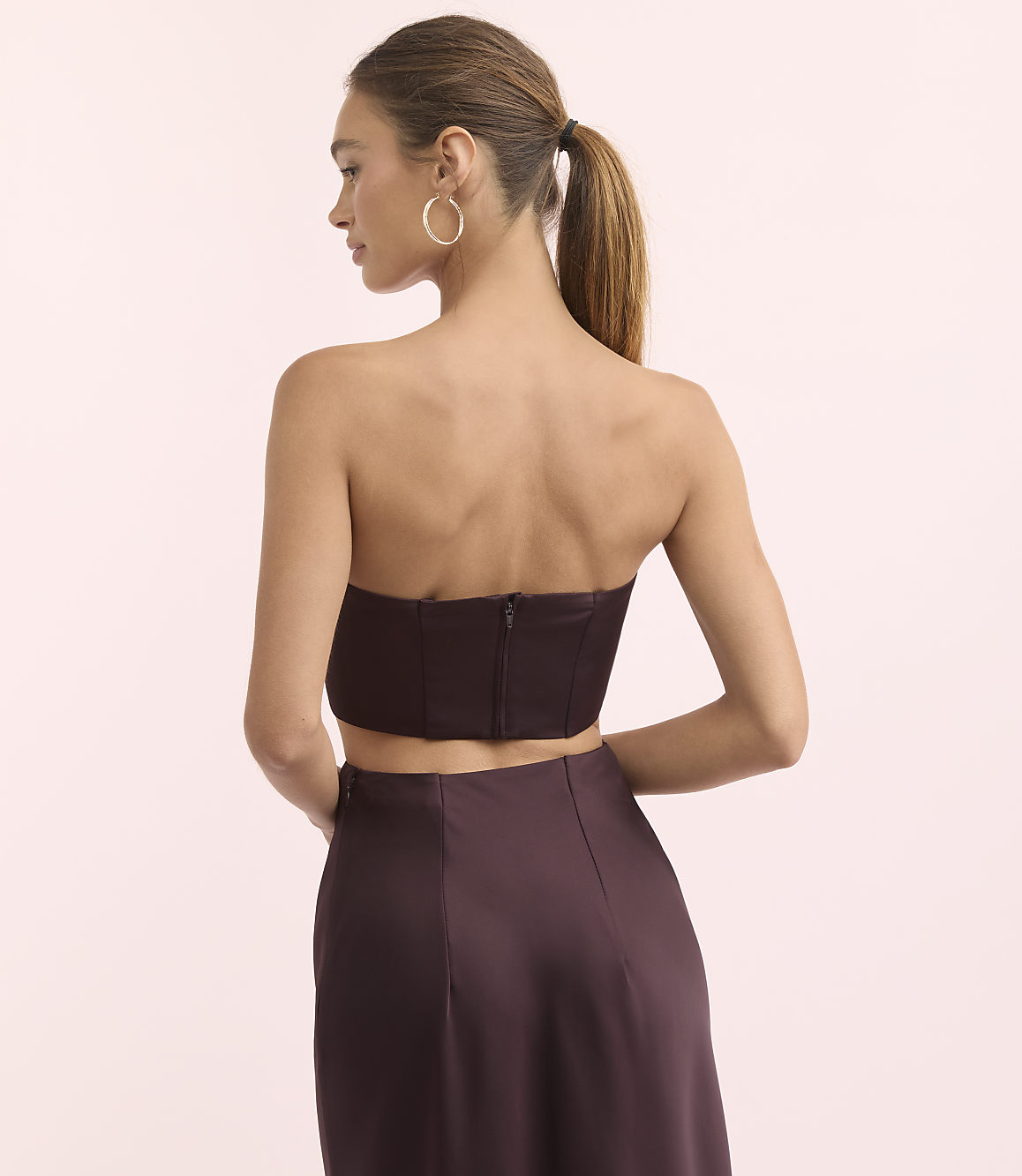 A woman in a strapless top and skirt stands against a plain pink background.