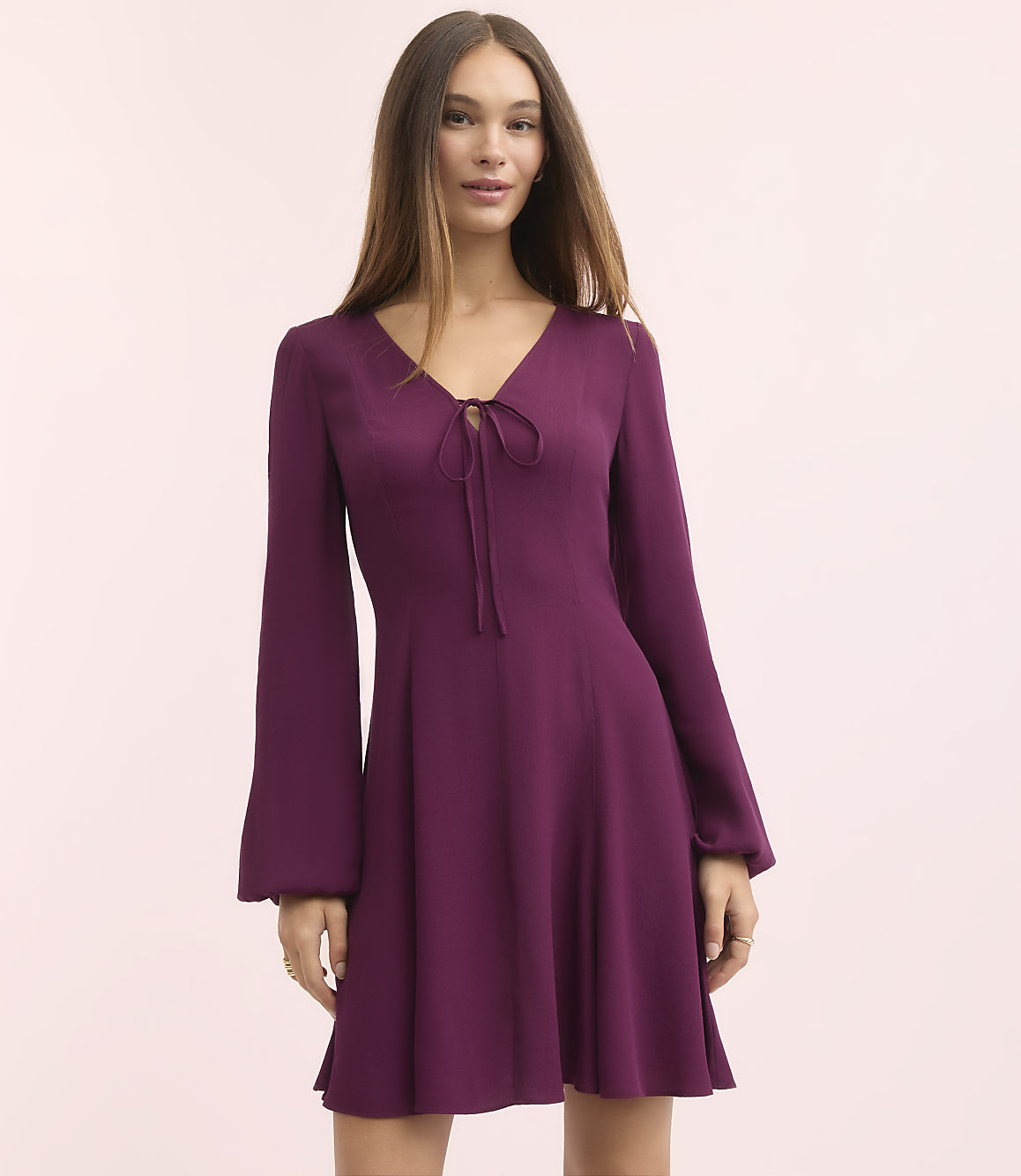 A young woman in a purple dress with bell sleeves stands against a soft pink background.