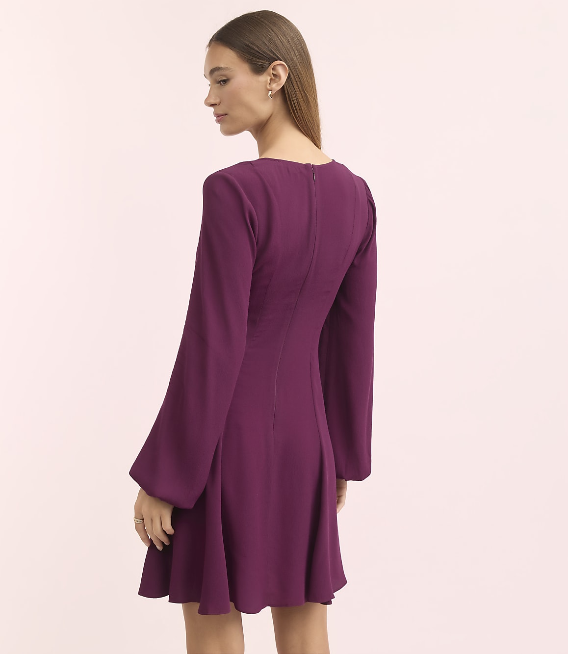 A woman in a purple dress with bell sleeves stands against a soft pink background.