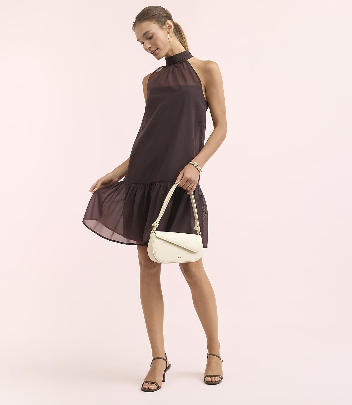 A woman in a dark purple halter dress and cream handbag stands against a soft pink background.