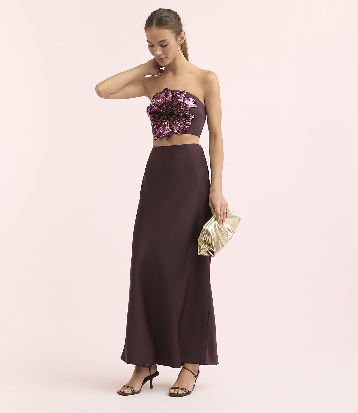 A woman in a strapless floral top and a long skirt poses with a gold clutch against a soft pink background.
