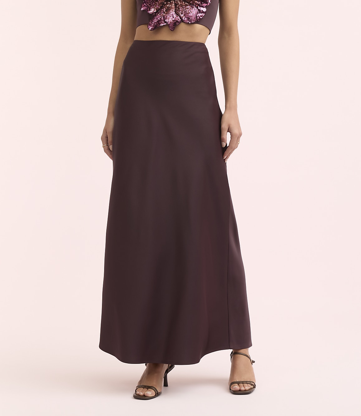 A woman in a long, dark purple skirt and a sequined top stands against a soft pink background.