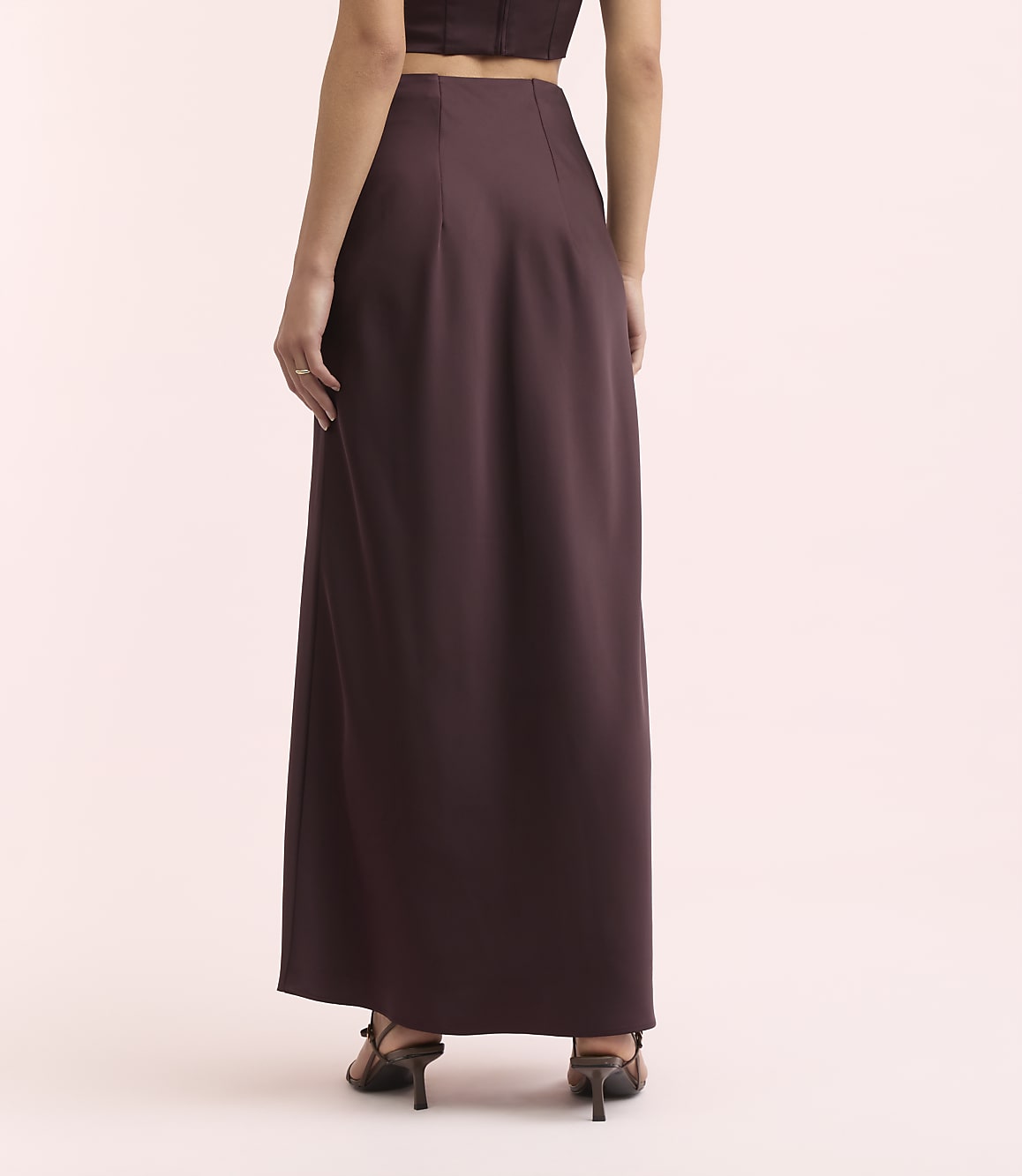 A person wearing a dark purple maxi skirt and heels stands against a soft pink background.