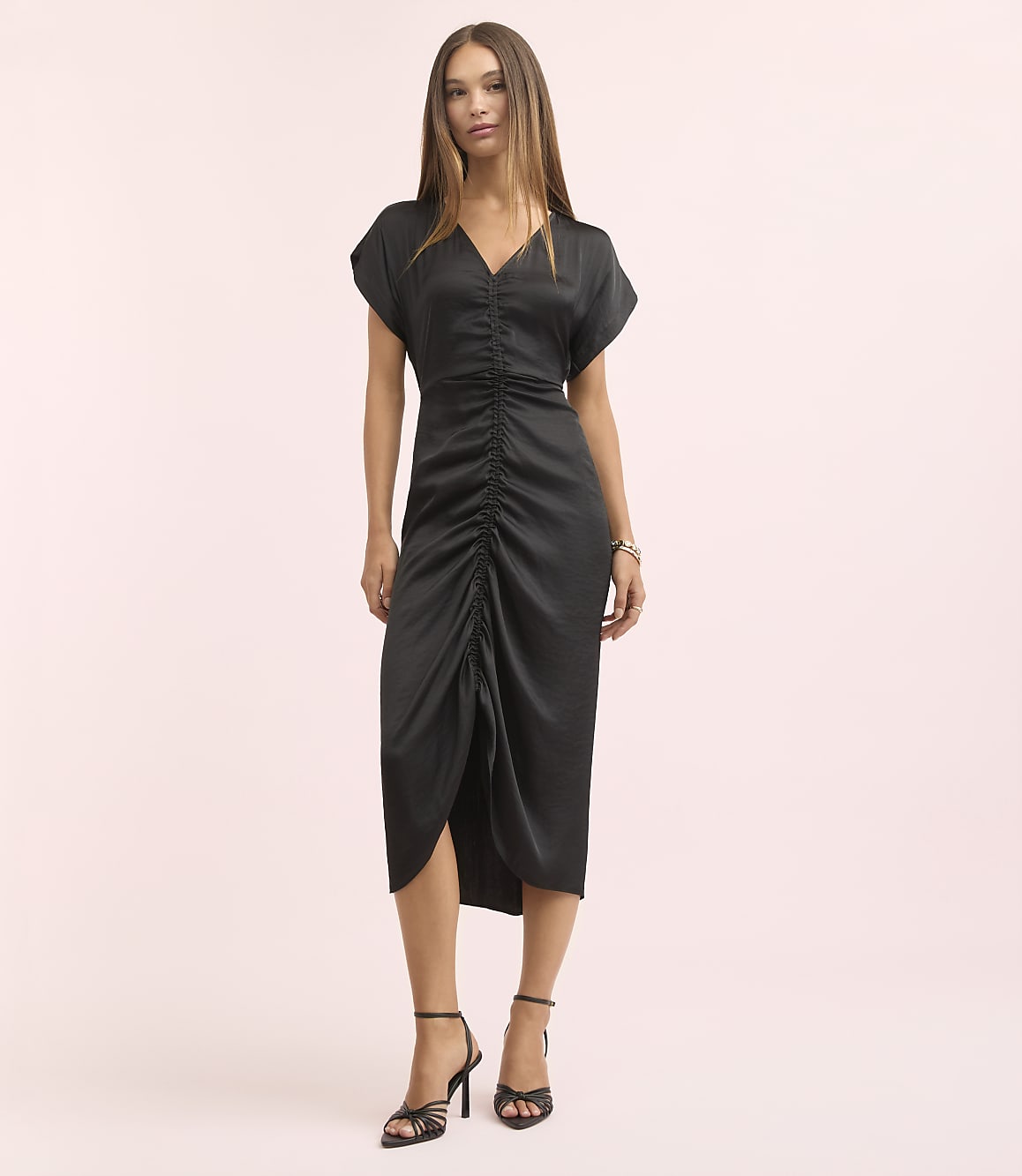 A woman in a black ruched dress and strappy heels stands against a soft pink background.
