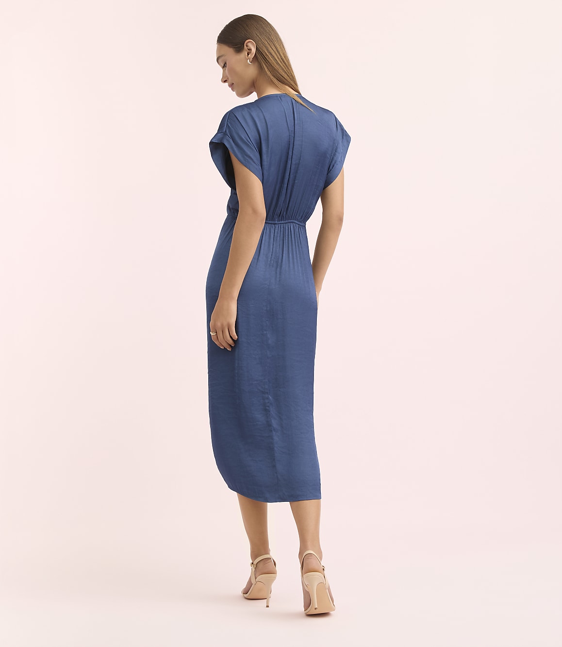A woman in a blue dress stands against a soft pink background.