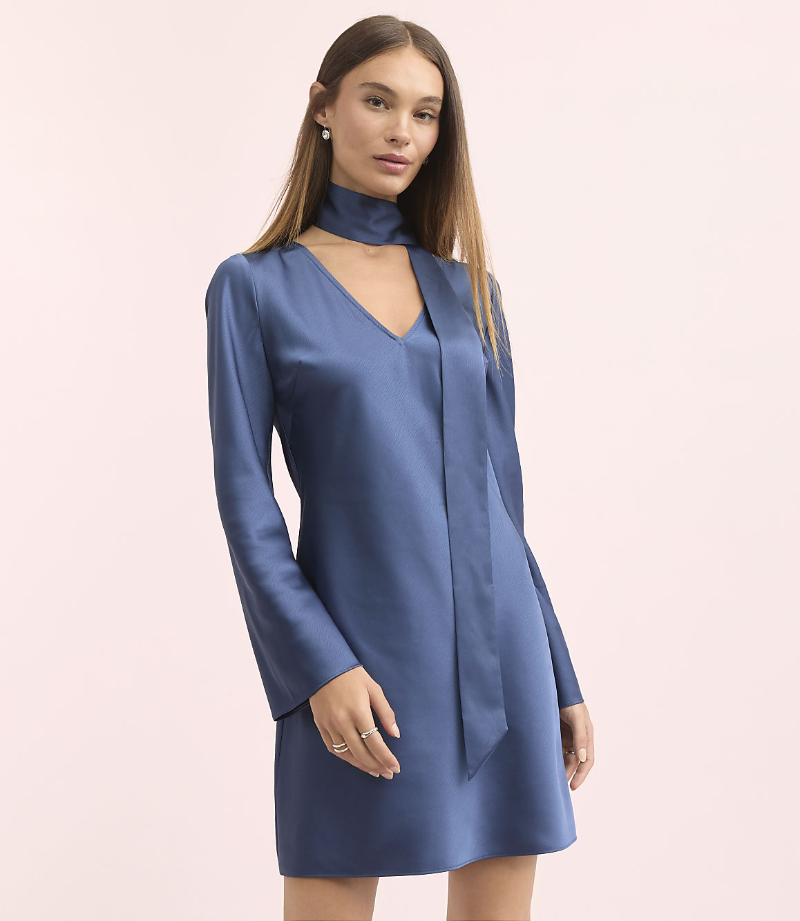 A woman in a blue satin dress with bell sleeves and a scarf neckline stands against a soft pink background.