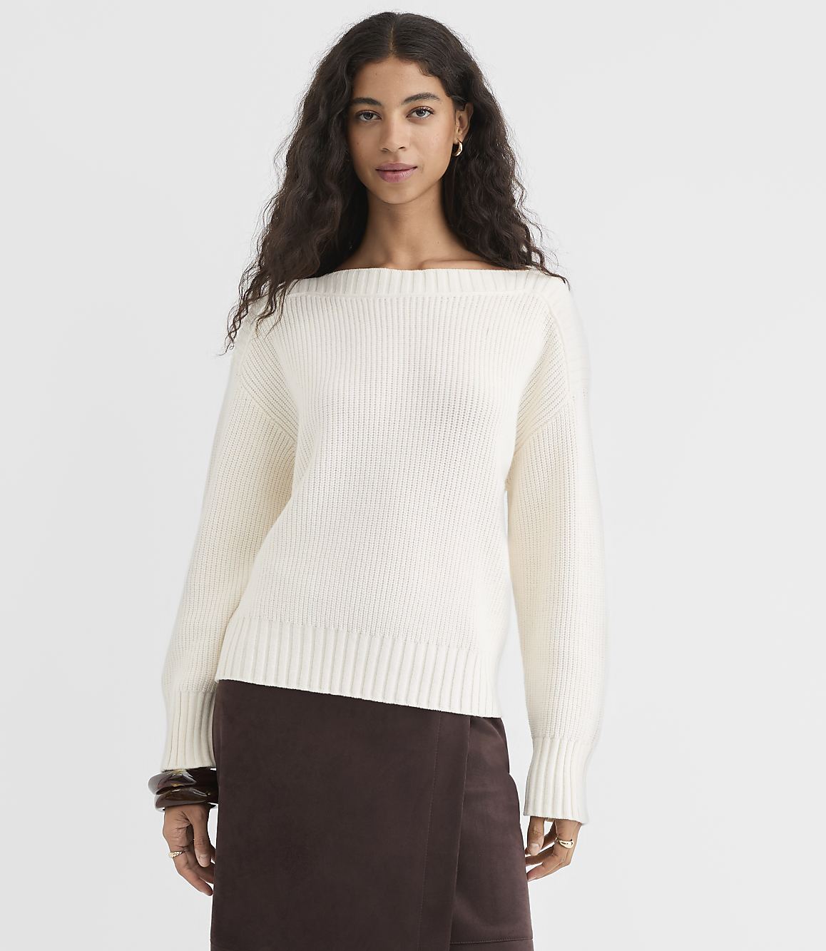 A woman in a cream-colored boatneck sweater and brown skirt stands against a plain white background.
