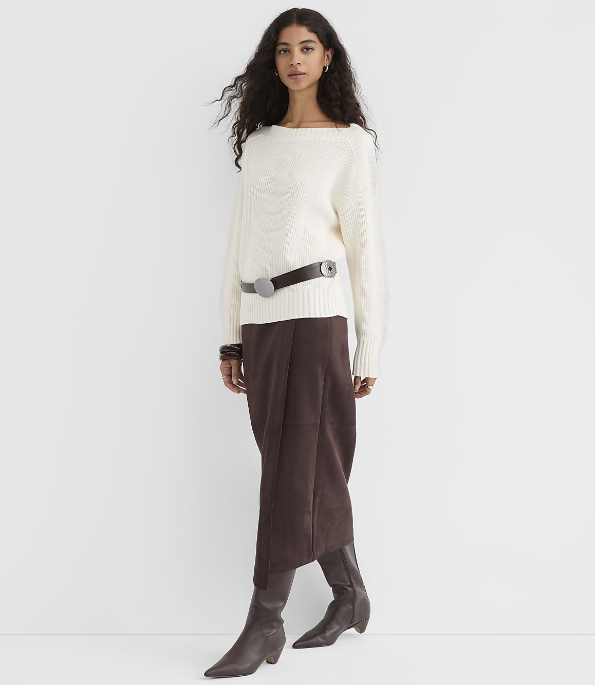 A woman in a cream sweater and brown skirt stands against a plain white background.