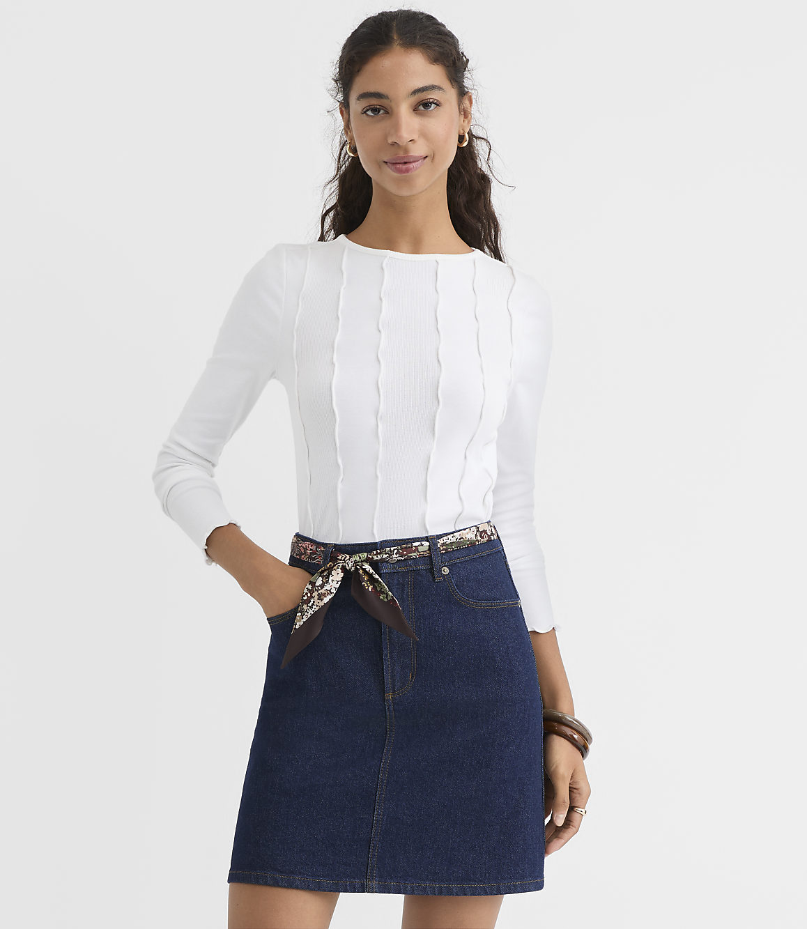 A woman in a white ribbed top and denim skirt stands against a plain white background.