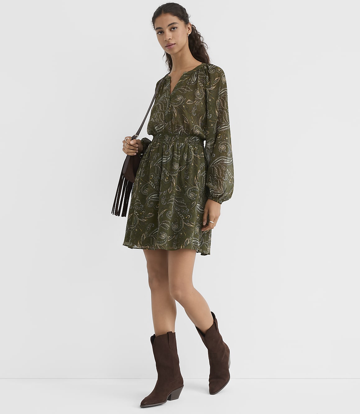 A woman in a green paisley dress and brown boots stands against a plain gray background, carrying a fringed brown bag.
