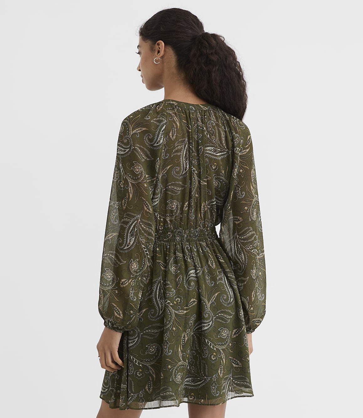 A woman in a green paisley dress with long sleeves stands against a plain, light background.
