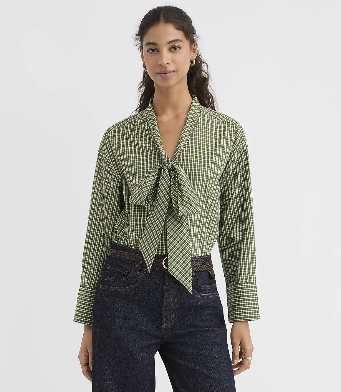 A woman in a green checkered blouse with a bow tie stands against a plain gray background.