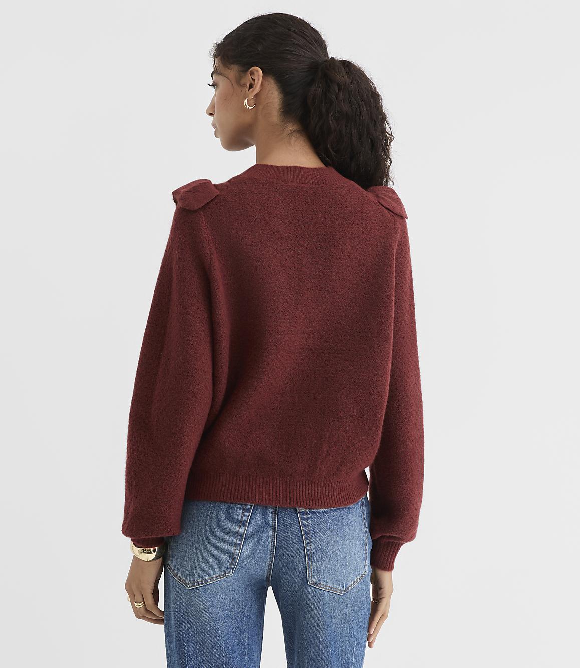 A woman in a maroon sweater and blue jeans stands against a plain white background.