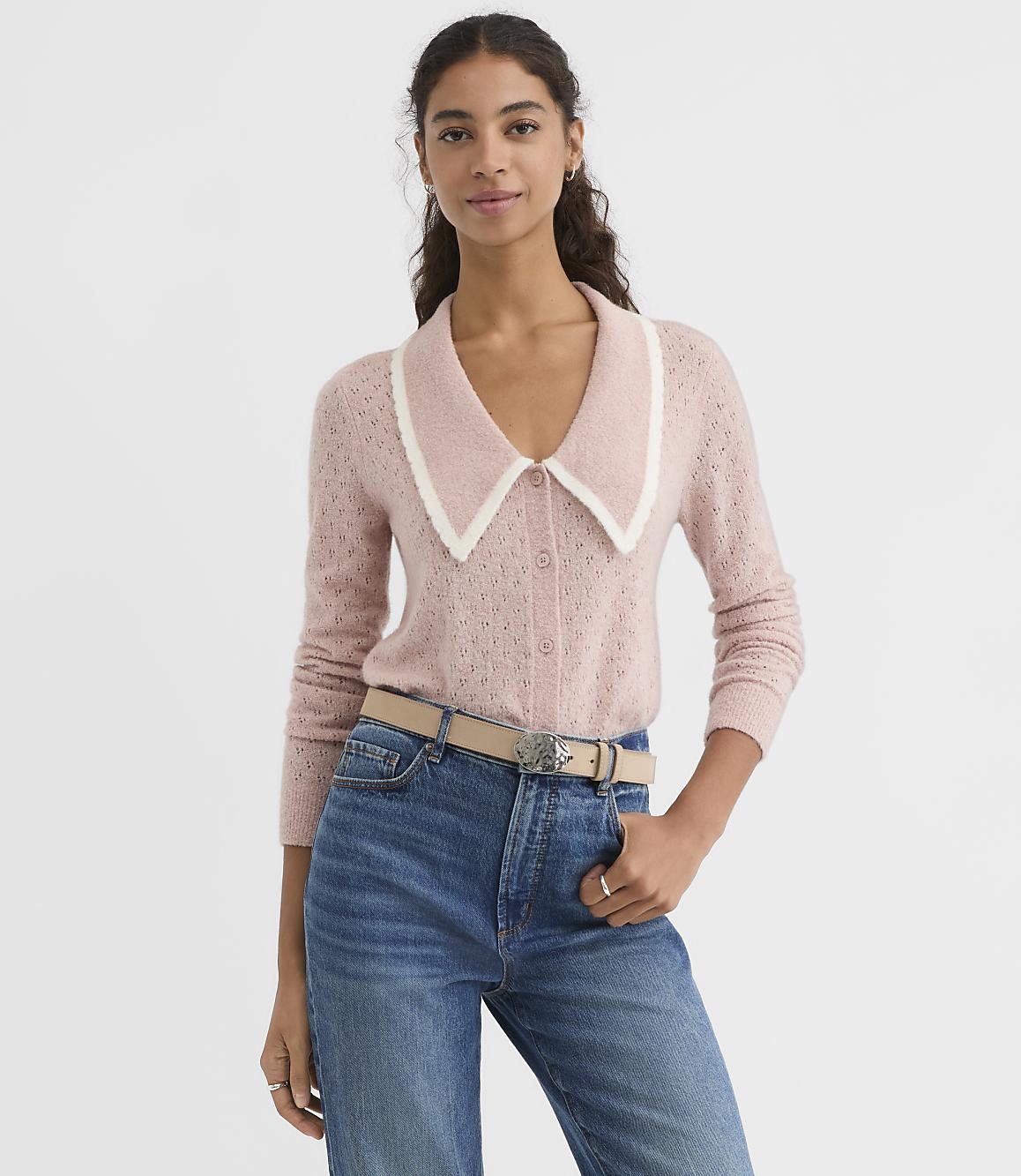 A woman in a pink cardigan with a white collar stands against a plain white background.