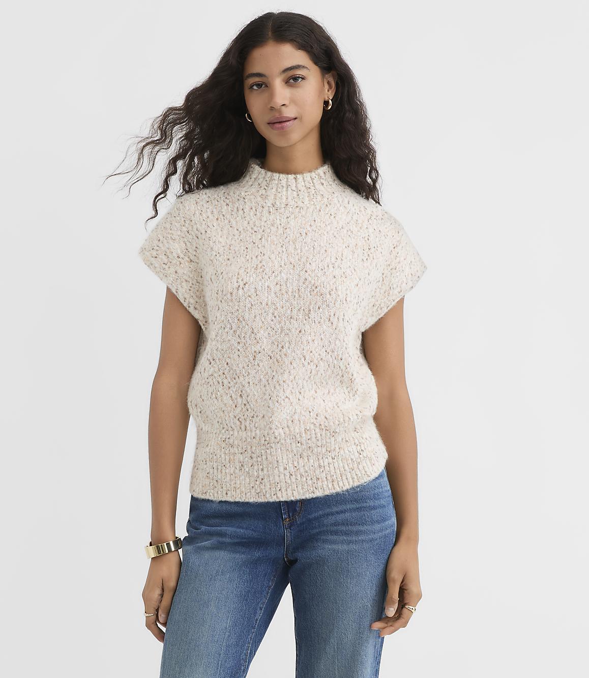 A woman in a speckled knit sweater and blue jeans stands against a plain white background.