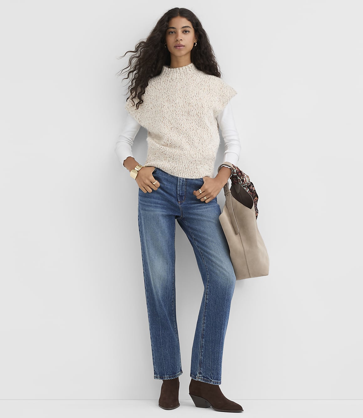 A woman in a cream speckled sweater vest and blue jeans stands against a plain white background, holding a tan tote bag.