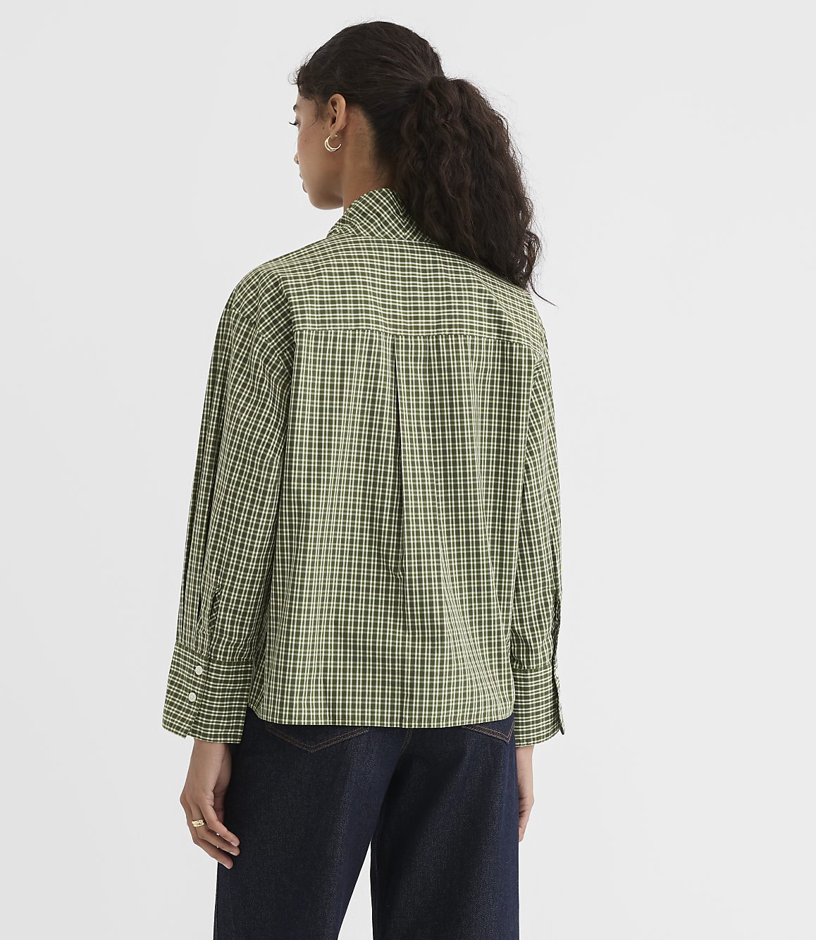 A woman in a green checkered shirt and dark jeans stands against a plain white background.