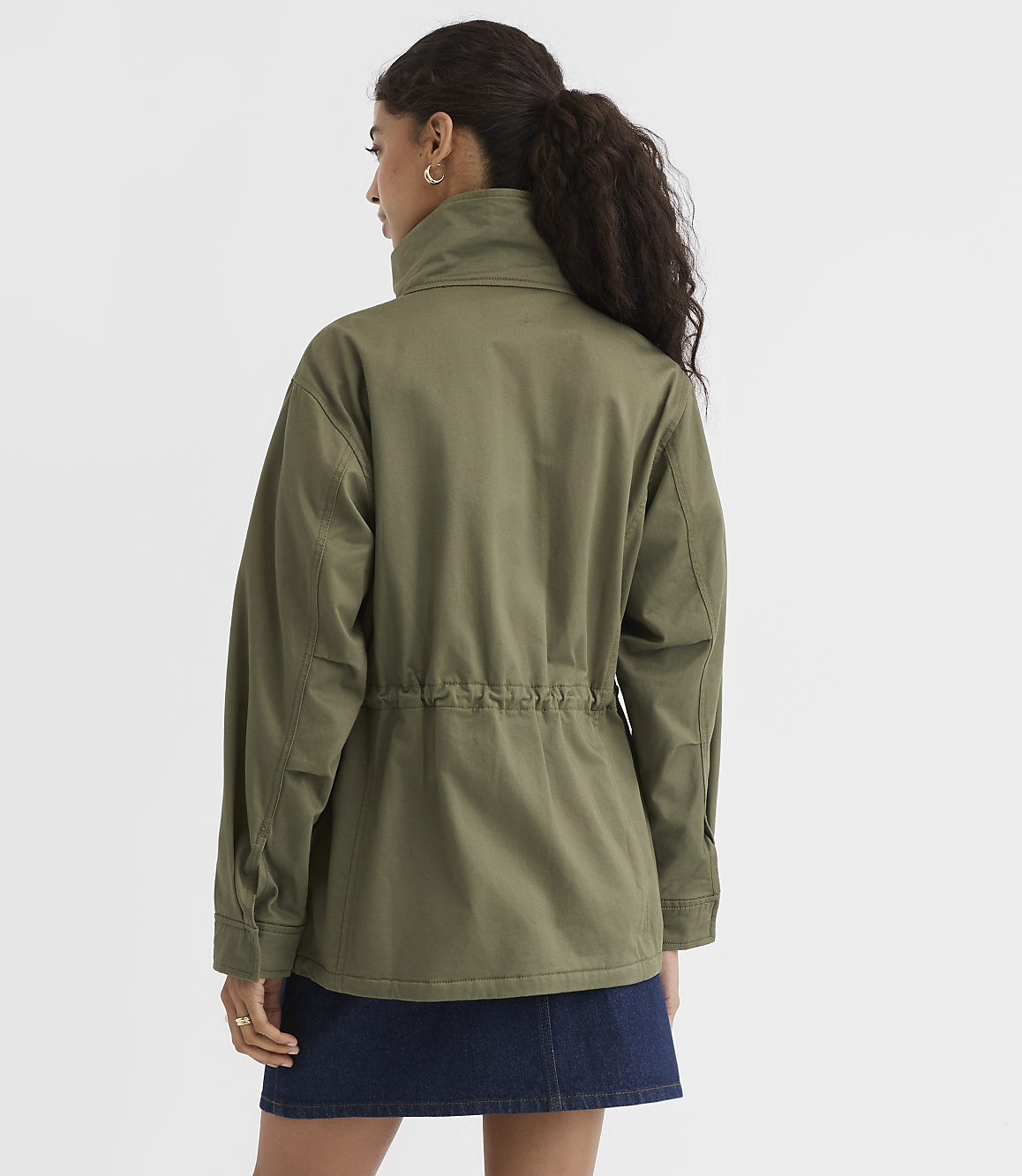 A woman in a green jacket and denim skirt stands against a plain white background.