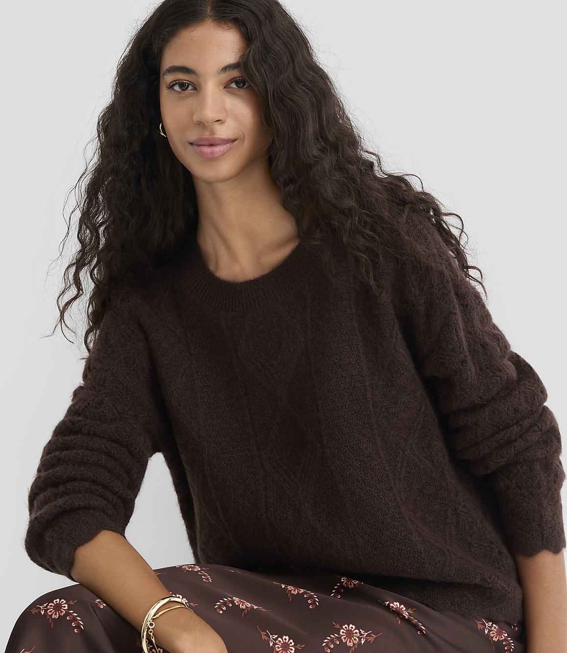 A woman with long, dark, wavy hair wears a dark brown, textured sweater and a patterned skirt, with a plain gray background.