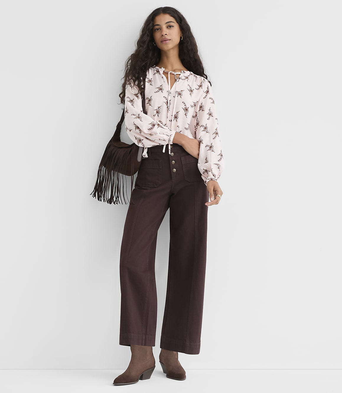A woman in a patterned blouse and wide-leg pants stands against a plain, light gray background.