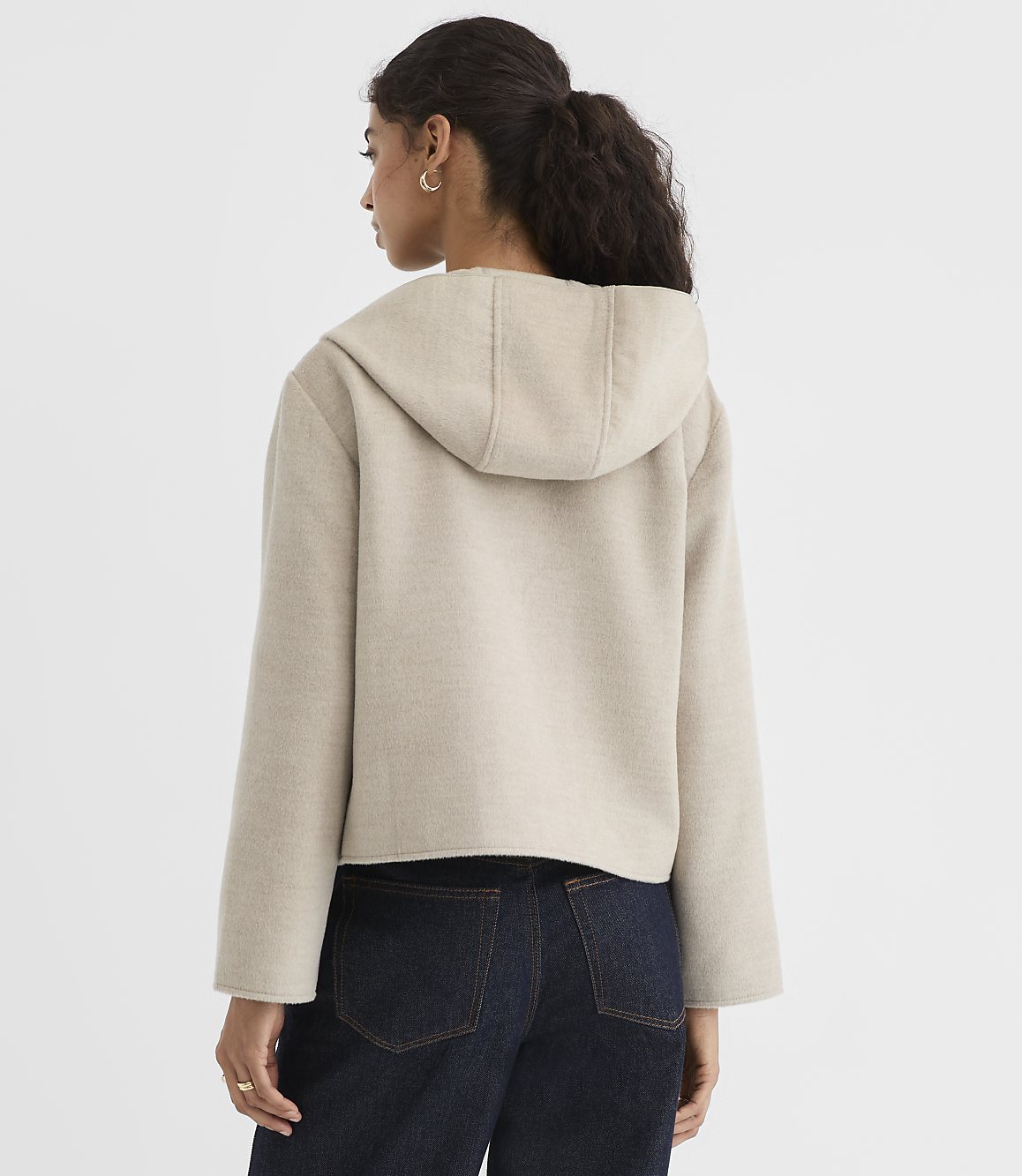 A woman in a beige hooded jacket and dark jeans stands against a plain white background.