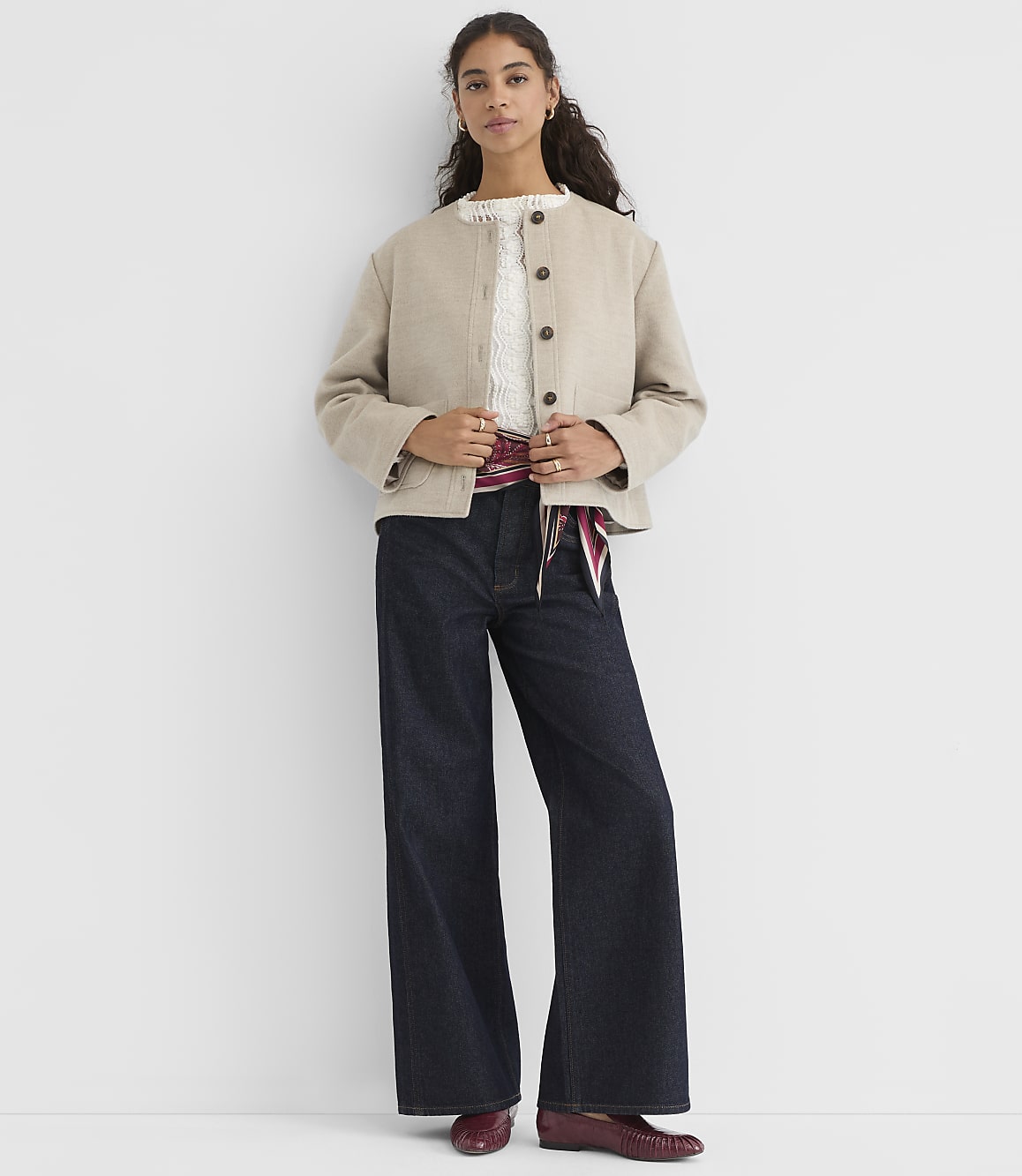 A woman in a beige jacket and dark wide-leg jeans stands against a plain white background.