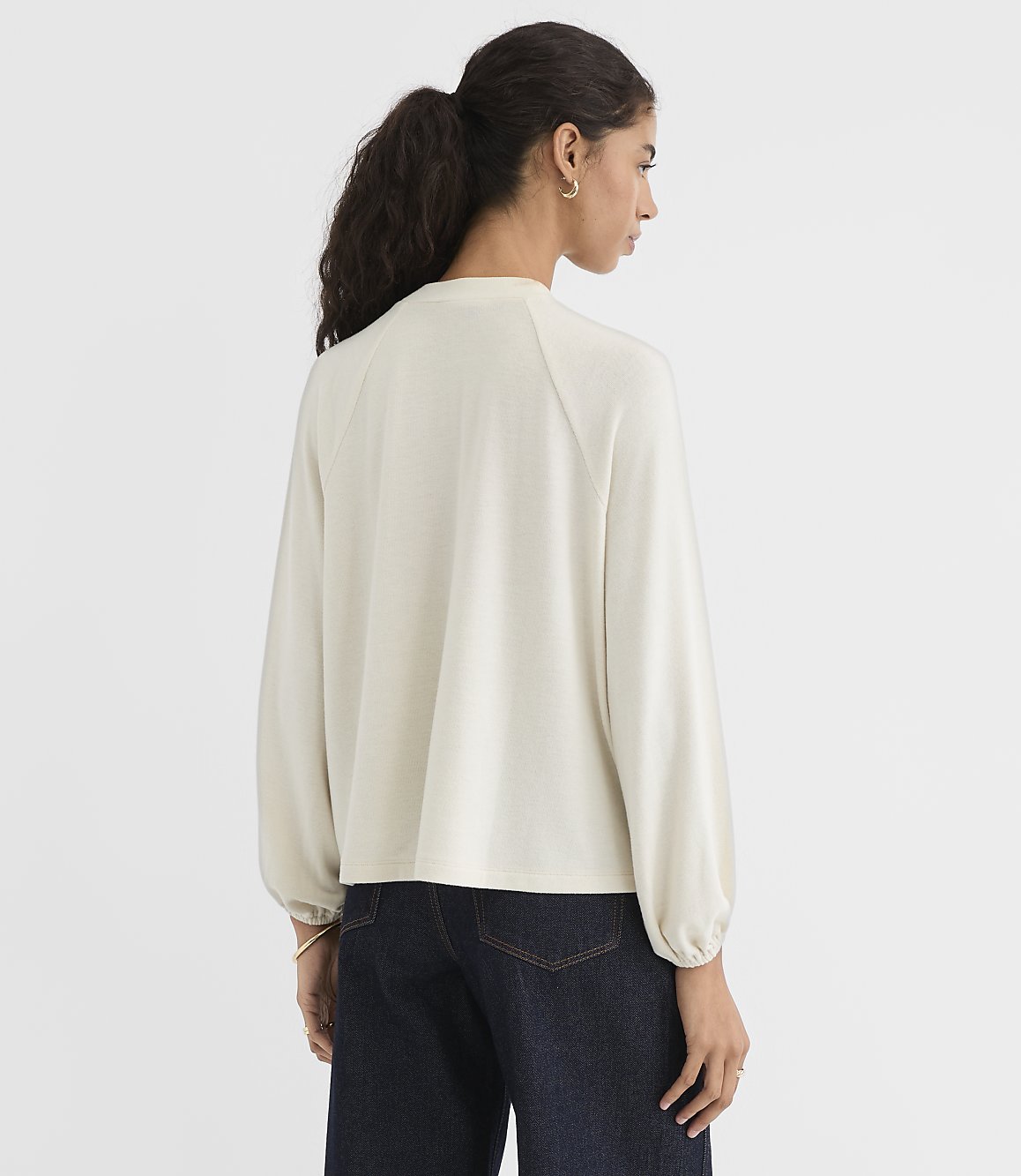 A woman in a cream-colored top and dark jeans stands against a plain white background.