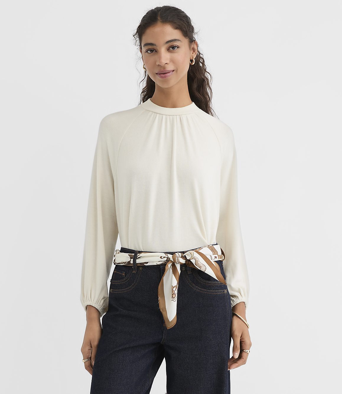A woman in a cream-colored mock-neck top and dark jeans stands against a plain white background, with a patterned scarf tied around her waist.