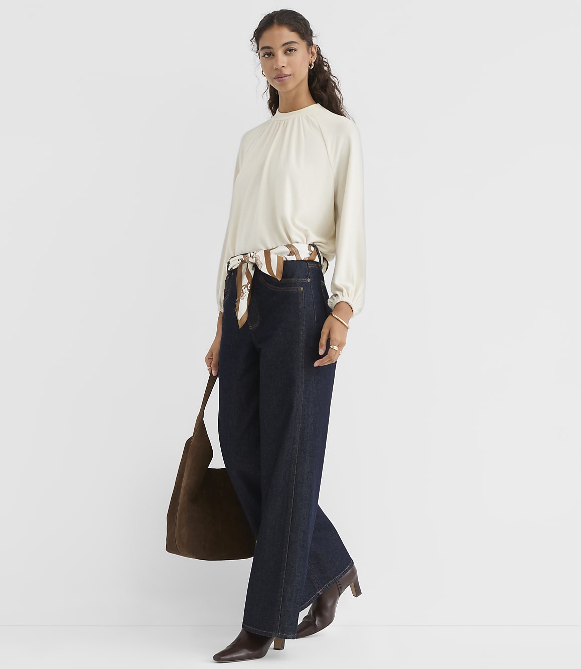A woman in a cream-colored blouse and dark wide-leg jeans stands against a plain white background, holding a brown suede tote bag.