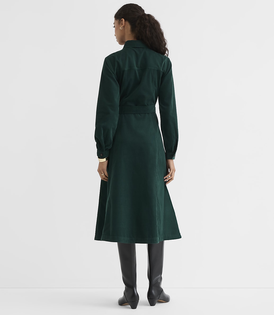 A woman in a green corduroy dress and black boots stands against a plain white background.