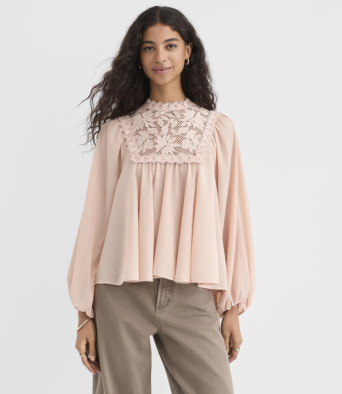 A woman in a light pink, lace-trimmed blouse and brown pants stands against a plain white background.