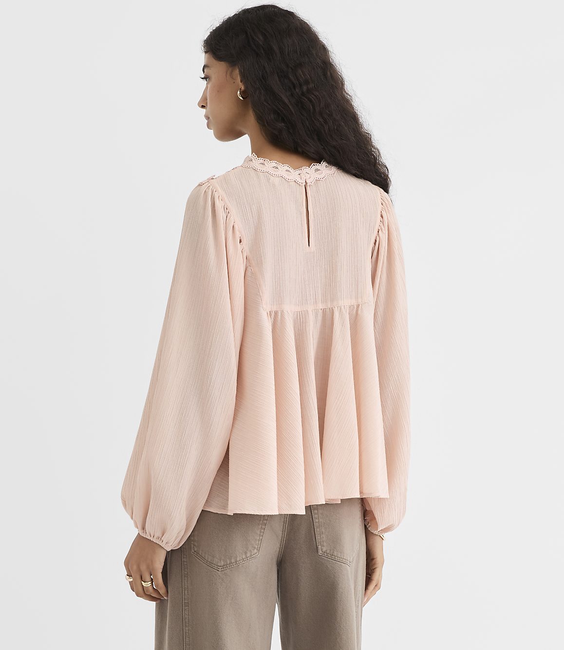 A woman in a pale pink blouse and brown pants stands against a plain, light-colored background.