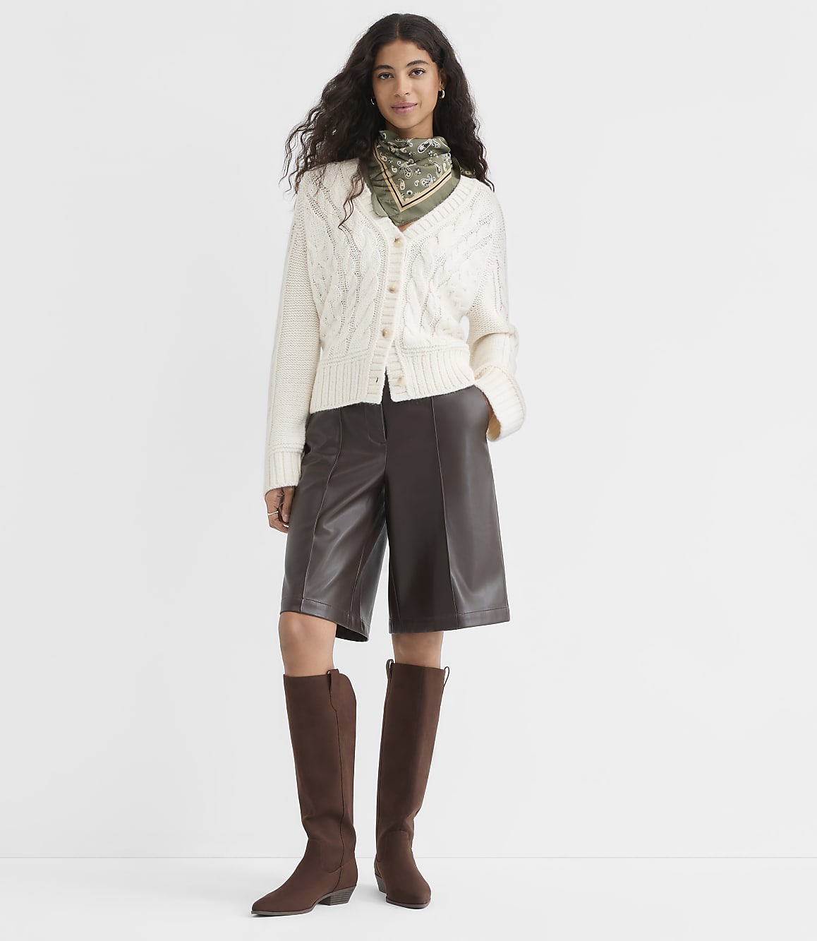 A woman in a cream cable-knit cardigan, brown leather culottes, and brown boots stands against a plain white background.