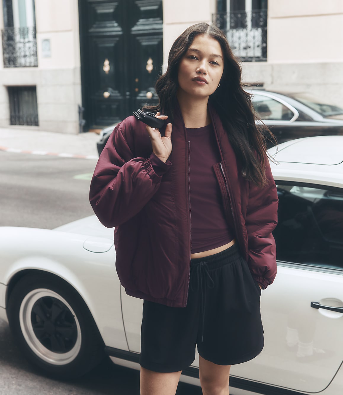 A young woman in a burgundy bomber jacket and black shorts stands in front of a white car on a city street with a large building in the background.
