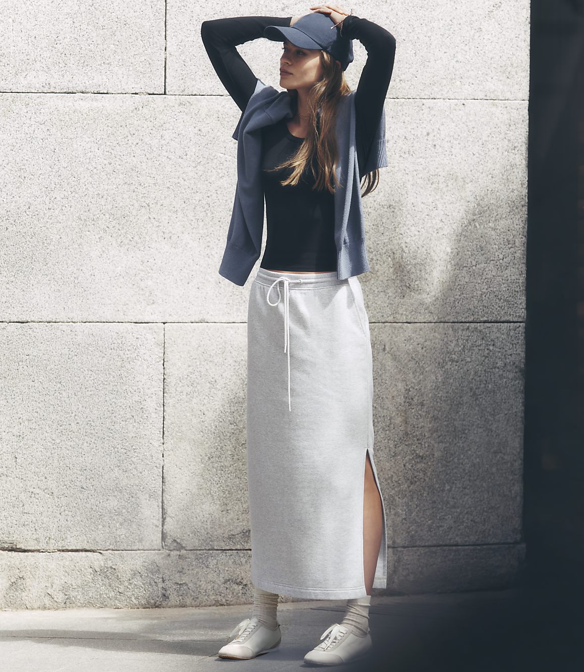 A woman in a black top, grey skirt, and blue cap stands against a textured stone wall.