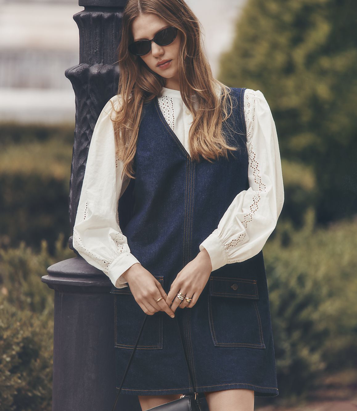 A young woman in a denim dress and white blouse leans against a lamppost with a blurred park in the background.