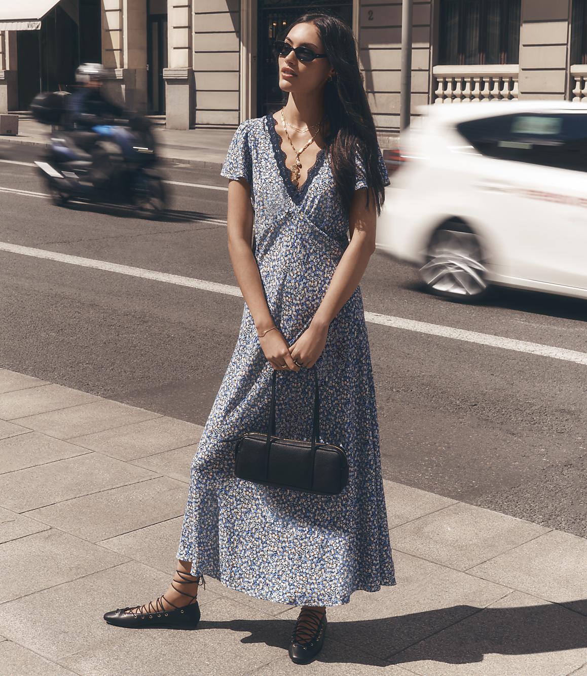 A woman in a floral dress stands on a sidewalk with a blurred motorcycle and car passing by in the background.