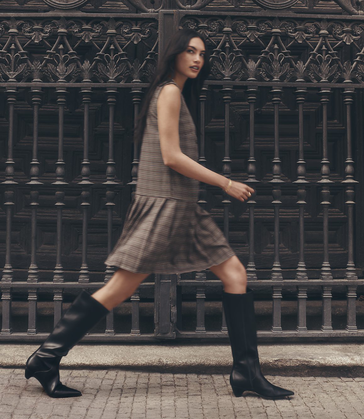 A woman in a plaid dress and black boots walks in front of an ornate metal gate.