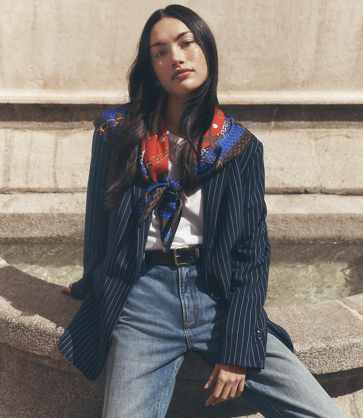 A young woman in a pinstripe blazer and jeans sits in front of a stone fountain.