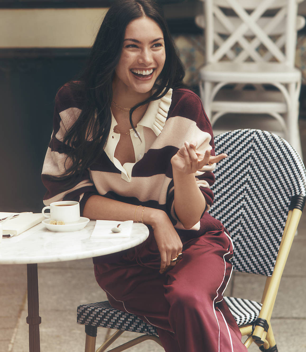 A laughing woman in a striped sweater and maroon pants sits at a small round table with a coffee cup, while a white chair is visible in the blurred background.