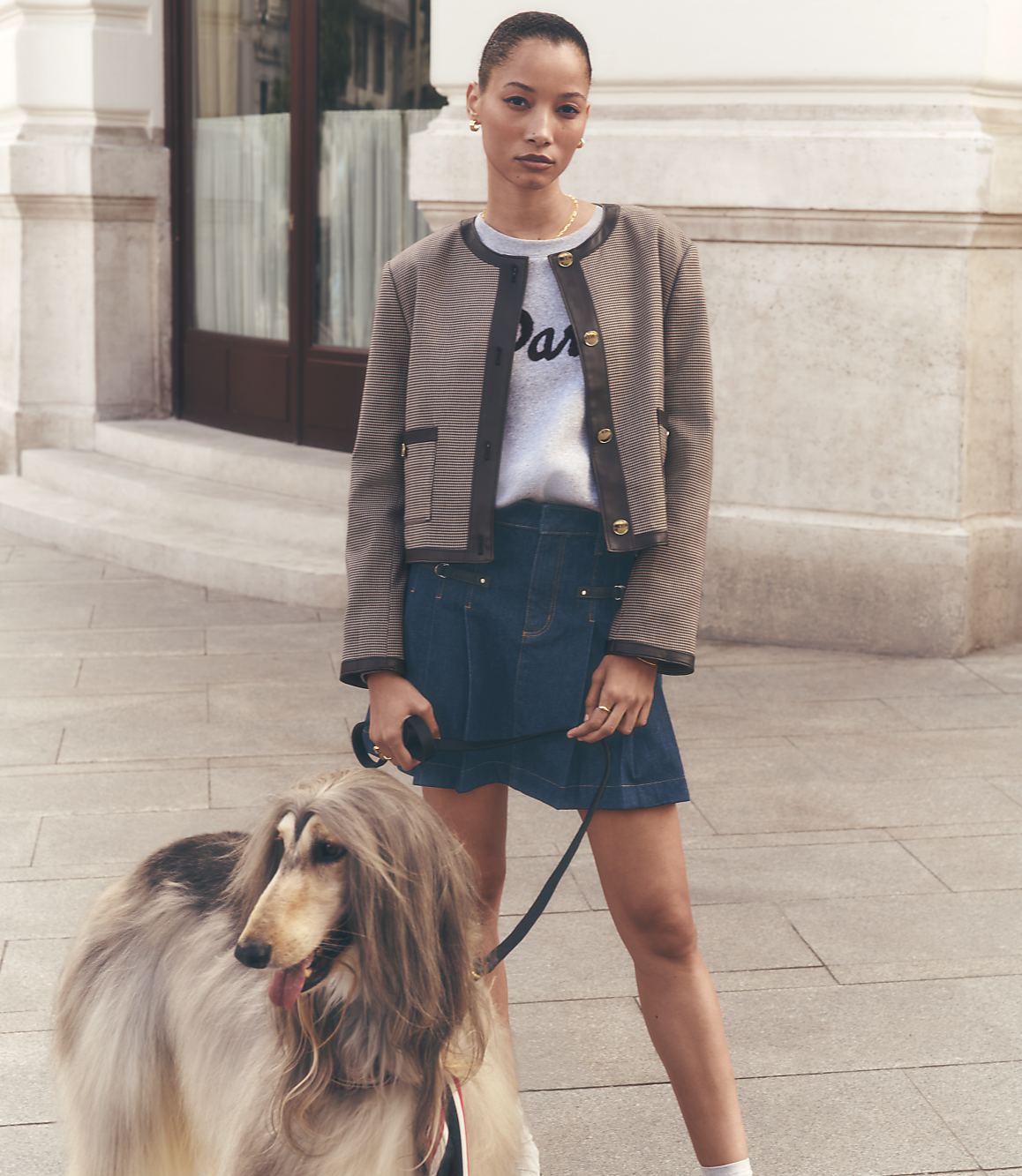 A woman in a stylish jacket and skirt stands with an Afghan hound on a sidewalk in front of a building.