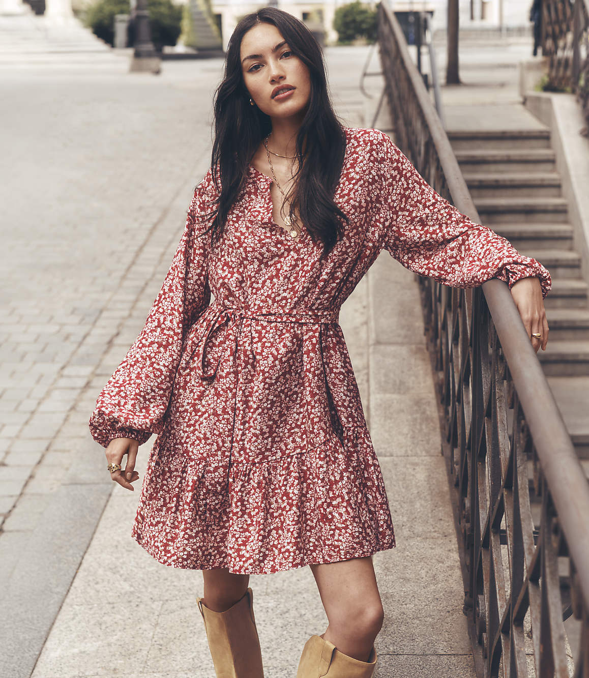 A woman in a floral dress and boots stands on a sidewalk next to a metal railing, with a blurred urban background.
