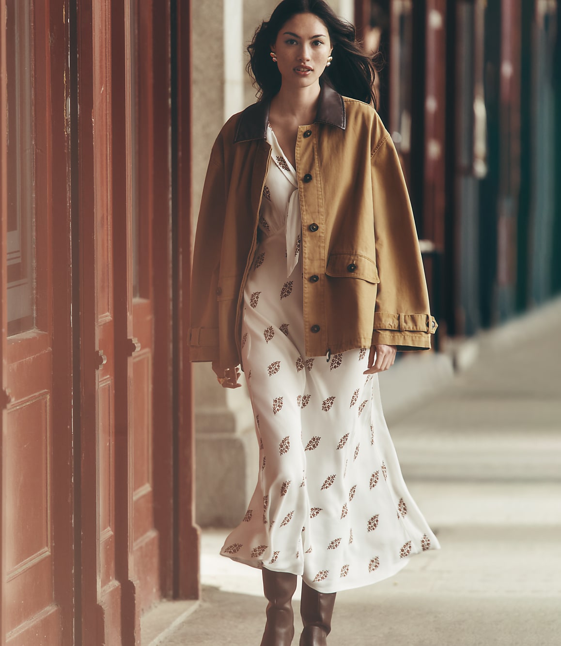 A woman in a patterned dress and brown jacket walks past a red wooden door with a blurred street in the background.