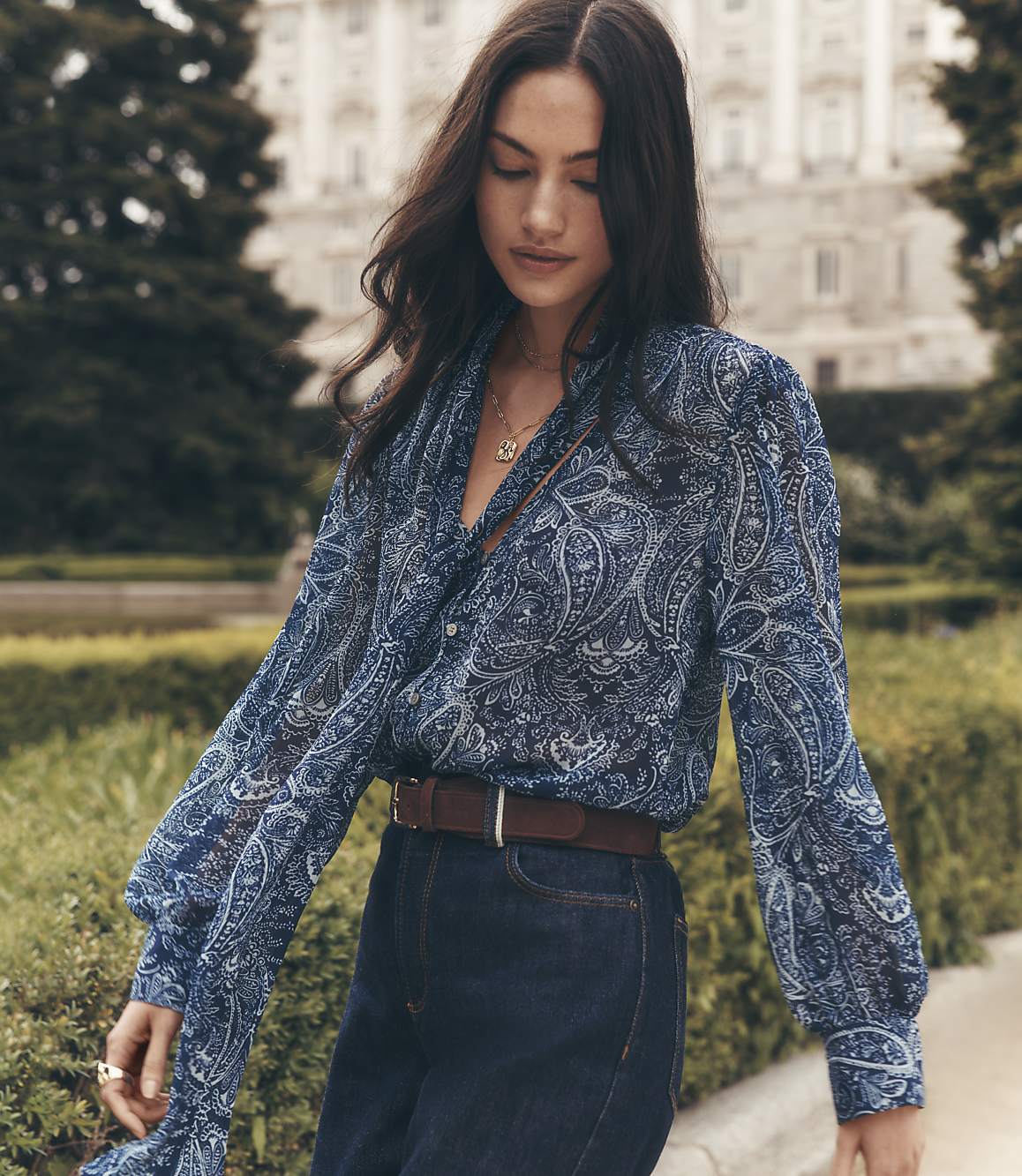 A woman in a blue paisley blouse and dark jeans stands in front of a grand building with trees.