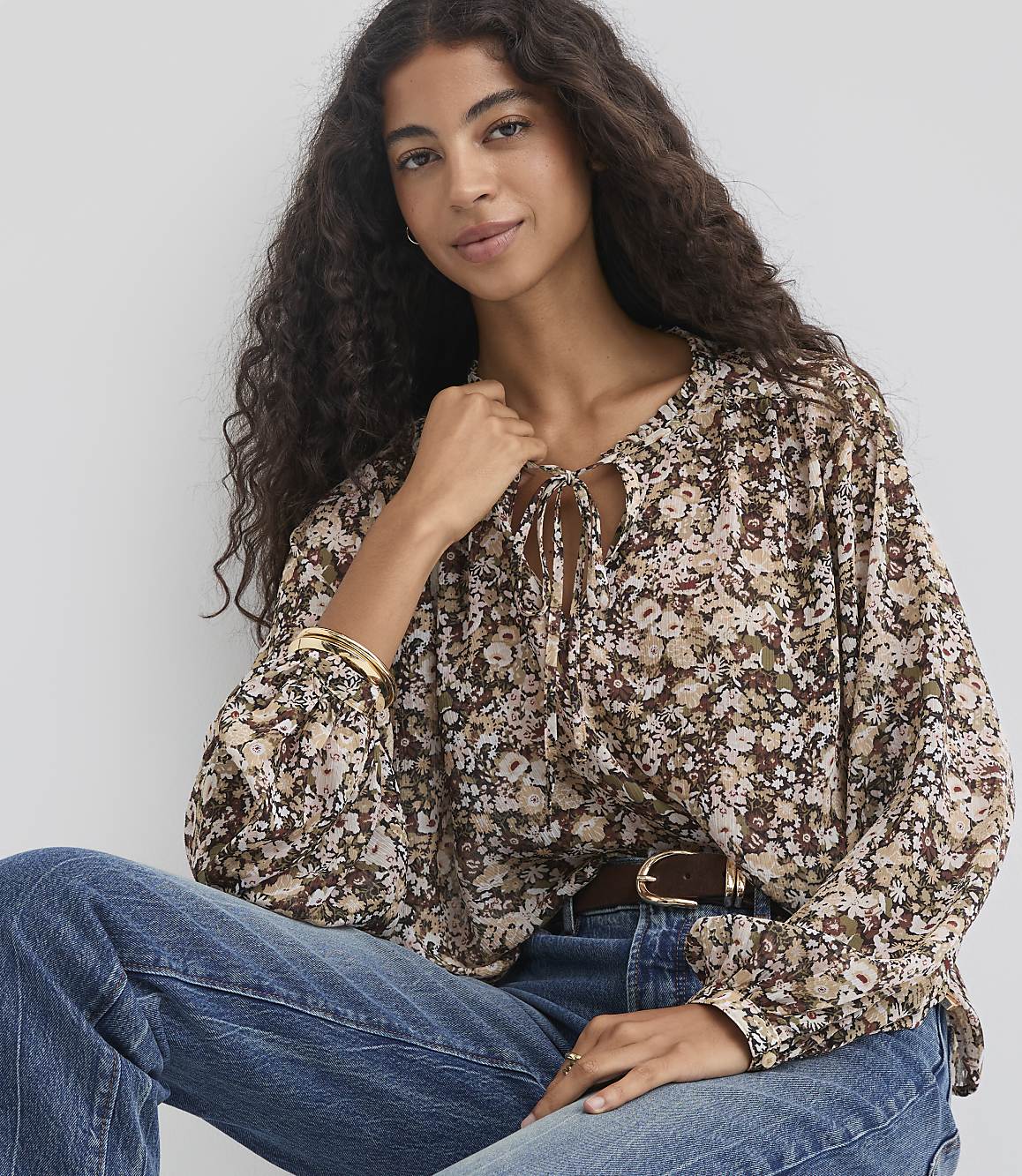A woman with long, dark, curly hair wears a floral print blouse and blue jeans, seated against a plain, light-colored background.